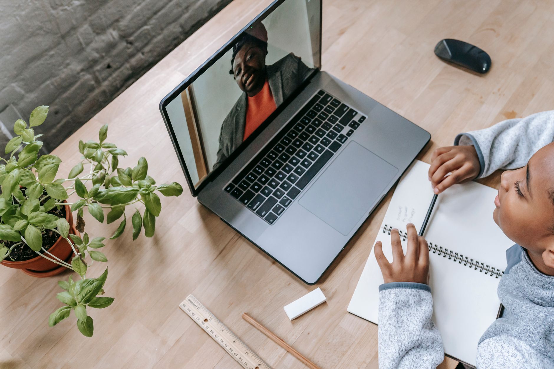 Top-down view of a desk with a laptop showing a tutor on a video call, a notebook and a plant